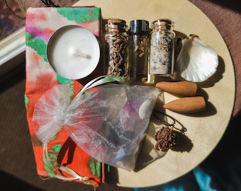 May include: A wooden tray with a variety of items for a spiritual ritual, including a white candle, a small bag of crystals, a seashell, incense sticks, and four small glass bottles with herbs and liquids.