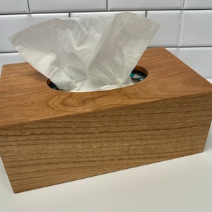 May include: A wooden tissue box cover with a natural wood finish. The box has a rectangular shape with a rounded opening for tissues. A white tissue is partially pulled out of the opening.