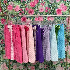 May include: A collection of eight girls' skirts in various colors, including pink, purple, and blue. The skirts are made of a soft, textured fabric and have a gathered design. Each skirt has a ribbon tied around the waist.