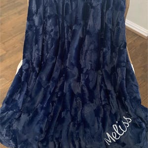 May include: Navy blue faux fur blanket draped over a chair. The blanket has a textured surface and is embroidered with the name "Melissa" in white script. The blanket is a deep navy color.