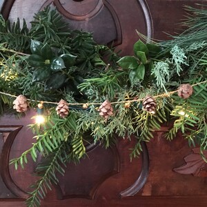 May include: A Christmas garland composed of various evergreen foliage, such as holly and pine. The garland is decorated with small pine cones, miniature gold bells, and warm white fairy lights, creating a festive holiday ambiance. The garland is placed on a dark wooden surface.