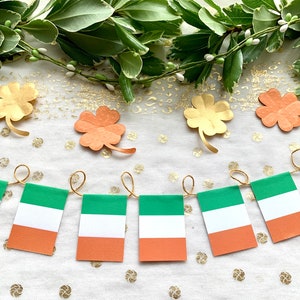 May include: A string of green, white, and orange Irish flags with gold string hanging from a white background with gold shamrocks.
