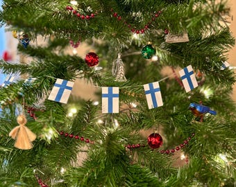 May include: A decorated Christmas tree with Finnish flag bunting, red bead garland, and various ornaments. The ornaments include red and green baubles, a blue bird, and a gold angel. The tree is lit with small white lights.