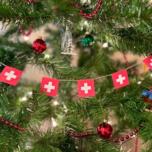 May include: A decorated Christmas tree with red and white Swiss flag bunting, red ornaments, and a small blue bird ornament. The tree is adorned with red bead garland and a silver trumpet ornament.