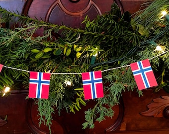 May include: A string of red and blue Norwegian flags hangs from a garland of green foliage with white Christmas lights.