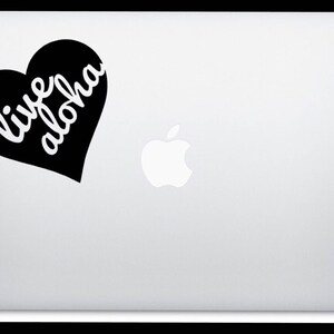 Op de afbeelding: Een zwarte hartvormige sticker met de woorden "live aloha" in witte letters is bevestigd op een zilveren laptop. Het Apple-logo is zichtbaar.