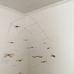 May include: A handmade mobile featuring multiple butterfly cutouts in various colors and patterns, suspended from a wire frame. The butterflies are in shades of white, beige, pink, and brown, creating a whimsical, decorative piece.
