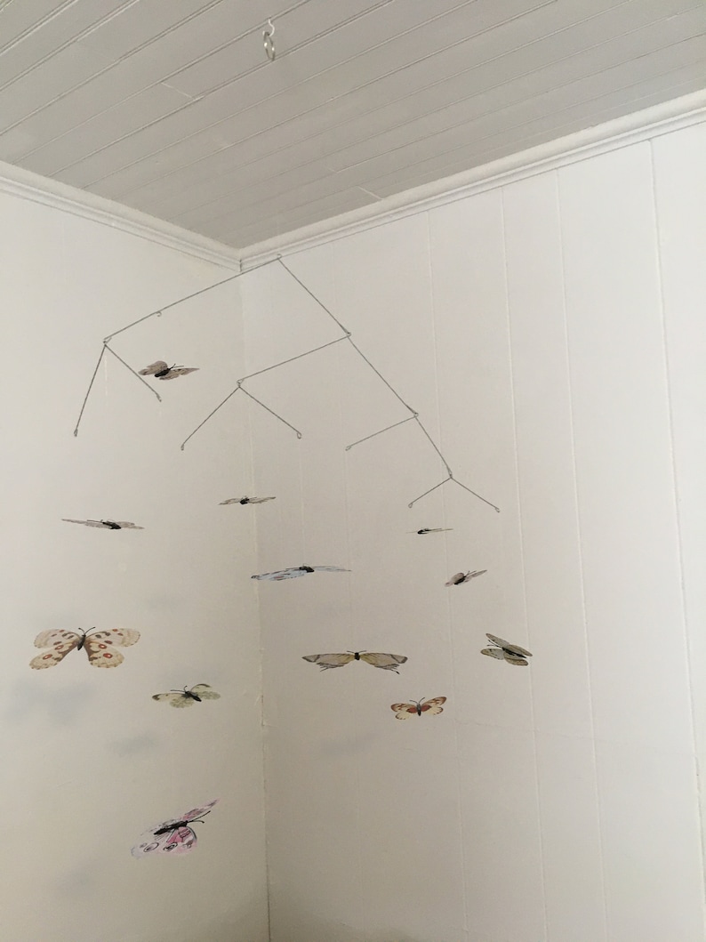 May include: A handmade mobile featuring multiple butterfly cutouts in various colors and patterns, suspended from a wire frame. The butterflies are in shades of white, beige, pink, and brown, creating a whimsical, decorative piece.
