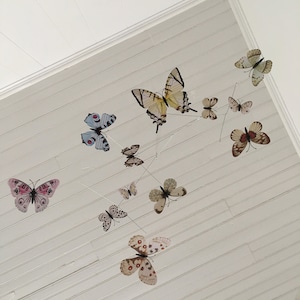May include: A collection of butterfly decorations suspended from a white, slatted ceiling. The butterflies vary in size, color, and pattern, including pink, blue, and brown. The arrangement creates a whimsical, airy aesthetic.