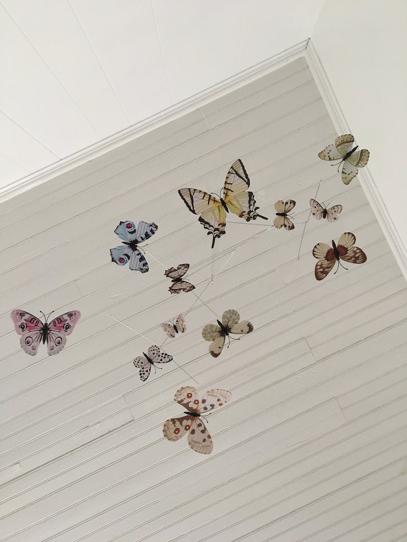 May include: A collection of butterfly decorations suspended from a white, slatted ceiling. The butterflies vary in size, color, and pattern, including pink, blue, and brown. The arrangement creates a whimsical, airy aesthetic.