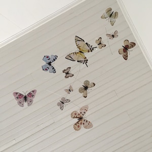 May include: A collection of decorative butterfly ornaments in various sizes and colors, suspended from thin wires against a white, slatted ceiling. The butterflies feature detailed wing patterns in shades of pink, brown, yellow, and blue.