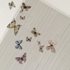 May include: A collection of decorative butterfly ornaments in various sizes and colors, suspended from the ceiling. The butterflies feature detailed wing patterns in shades of pink, brown, white, and blue, creating a whimsical, airy display.