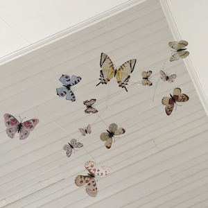 May include: A collection of decorative butterfly ornaments in various sizes and colors, suspended from thin strings against a white, slatted ceiling. The butterflies feature detailed wing patterns in shades of pink, blue, brown, and cream.