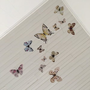 May include: A mobile featuring various butterfly designs in shades of yellow, brown, pink, and blue. The butterflies are suspended from thin wires against a white, slatted ceiling, creating a whimsical, decorative accent.