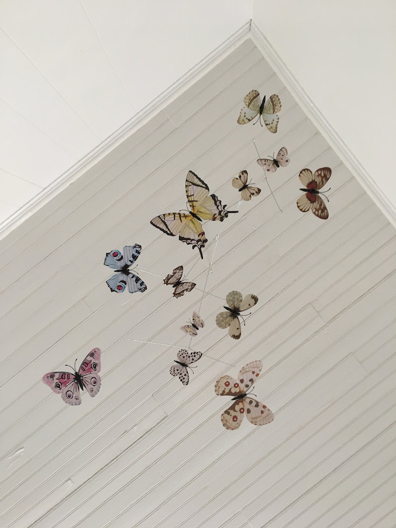 May include: A mobile featuring various butterfly designs in shades of yellow, brown, pink, and blue. The butterflies are suspended from thin wires against a white, slatted ceiling, creating a whimsical, decorative accent.