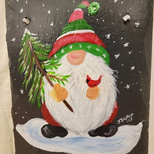 May include: A hand-painted slate sign with a gnome holding a small pine tree and a cardinal. The gnome is wearing a green hat with a red and white stripe and a white beard. The sign says "Welcome" in white paint and has a snowy background.