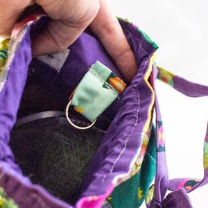 K&ouml;nnte beinhalten: Eine lila Stofftasche mit einem bunten Muster. Die Tasche hat einen Rei&szlig;verschluss und eine kleine Schlaufe innen. Die Schlaufe ist aus hellgr&uuml;nem Stoff mit einem silbernen Ring.