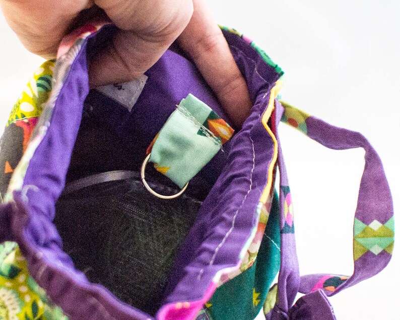K&ouml;nnte beinhalten: Eine lila Stofftasche mit einem bunten Muster. Die Tasche hat einen Rei&szlig;verschluss und eine kleine Schlaufe innen. Die Schlaufe ist aus hellgr&uuml;nem Stoff mit einem silbernen Ring.