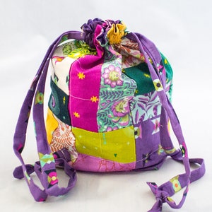 Könnte beinhalten: Eine violette Kordeltasche aus Patchworkstoff mit verschiedenen Farben und Mustern, darunter Pink, Grün, Gelb und Orange. Die Tasche hat einen gerafften oberen Rand und lange violette Kordeln.