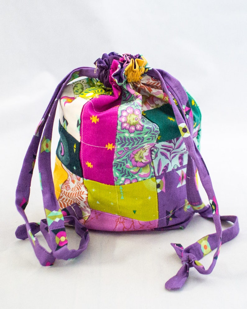 K&ouml;nnte beinhalten: Eine violette Kordeltasche aus Patchworkstoff mit verschiedenen Farben und Mustern, darunter Pink, Gr&uuml;n, Gelb und Orange. Die Tasche hat einen gerafften oberen Rand und lange violette Kordeln.