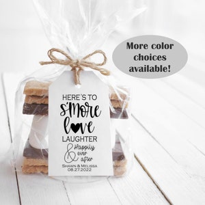 May include: A white tag with black text that reads "Here's to s'more love Laughter Happily ever after Shawn & Melissa 08.27.2022" is tied to a clear plastic bag containing a s'more. The s'more is made of graham crackers, chocolate, and a marshmallow. The bag is tied with twine. The background is a white wooden surface. The text "More color choices available!" is written in black on a gray oval.
