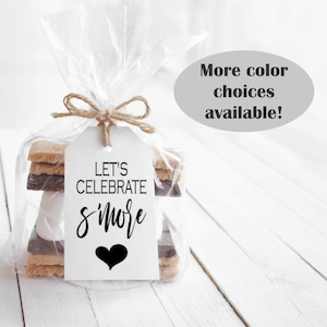 May include: A white gift tag with black text that reads "Let's Celebrate S'more" with a black heart. The tag is tied to a clear plastic bag containing a s'more with graham crackers, chocolate, and a marshmallow.