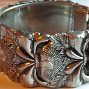 May include: Silver bracelet with a detailed floral design. The bracelet is made of multiple interlocking pieces that create a unique and intricate pattern.