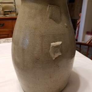 May include: A large, gray stoneware crock with a wide mouth and a rounded body. The crock has two small, irregular patches of glaze missing on the side.