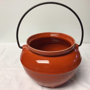 May include: A round, orange ceramic pot with a black metal handle. The pot has a glossy finish and a wide opening. The handle arches over the top of the pot, connecting on either side. The pot is likely for cooking or decorative purposes.