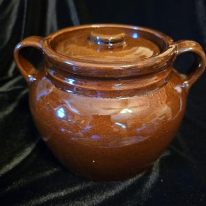 May include: A round, brown glazed ceramic pot with a matching lid and two side handles. The pot features a smooth, glossy finish and a wide, rounded body. The lid has a small, centered knob handle on top, and the overall design is simple.