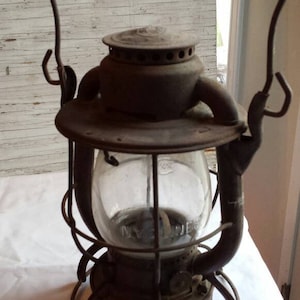 May include: An antique oil lamp with a glass globe and a metal cage. The lamp has a handle and is made of metal. The lamp is rusty and has a worn look.
