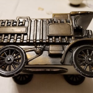 Peut inclure: Figurine de voiture ancienne de couleur bronze avec des roues noires. Le mod&egrave;le d&eacute;taill&eacute; comprend un radiateur, des phares et un capot incurv&eacute;. La voiture est un objet d&eacute;coratif, probablement un objet de collection ou un ornement de bureau.