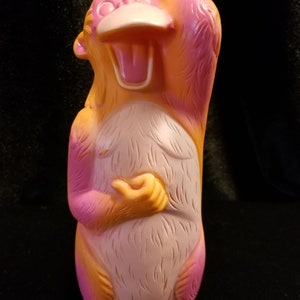 May include: A pink and orange rubber monkey toy with a wide open mouth and a hand on its head. The monkey is standing upright with its feet together.