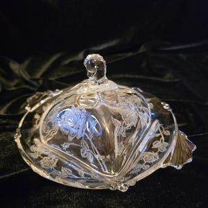 May include: A clear glass butter dish with a lid featuring an etched rose pattern. The lid has a small knob handle at the top, and the base has scalloped edges. The glass is transparent with white etched floral details throughout the surface.