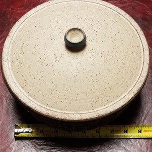 May include: A white ceramic casserole dish with a speckled glaze and a blue knob on the lid. The dish has a brown rim and is sitting on a red surface.