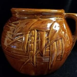 May include: A brown ceramic pitcher with a handle and a design of corn husks and ears of corn.