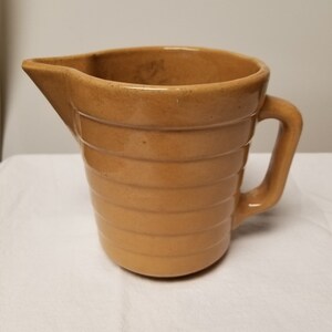 May include: A brown ceramic pitcher with a ribbed design and a handle. The pitcher has a slightly off-white color and a smooth, glazed finish.