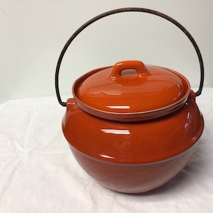 May include: A round, orange-glazed ceramic pot with a matching lid and a dark metal handle. The pot has a rounded body and a lid with a small handle. The handle arches over the top of the pot. The pot is sitting on a white surface.