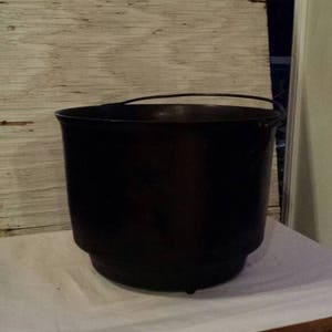 May include: A large black cast iron pot with a metal handle sits on a white tablecloth covered table with wooden legs.