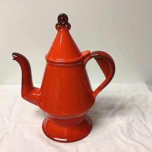 May include: A vibrant orange ceramic teapot with a conical lid and a curved spout. The teapot features a rounded base and a handle with a geometric design. The lid is topped with a decorative finial. The teapot is approximately 12 inches tall.