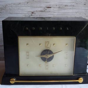 May include: A black vintage Admiral clock with a gold trim and a white face. The clock has a gold-colored hour and minute hand and a black second hand.
