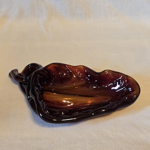 May include: A deep amber-colored glass dish shaped like a curled leaf. The piece features a translucent, organic form with smooth, undulating edges and a glossy finish that catches the light, creating varied tones of dark brown and golden orange.