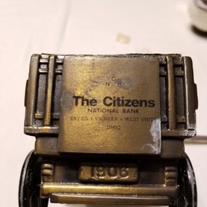 Peut inclure: Une voiture jouet vintage de couleur laiton avec des roues noires. La voiture porte l'inscription "The Citizens National Bank" et "1906". Il s'agit d'une r&eacute;plique d'un v&eacute;hicule du d&eacute;but du XXe si&egrave;cle.