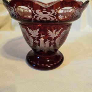 May include: A deep red glass bowl featuring a scalloped rim with cut-out details. The exterior surface is decorated with etched white designs depicting a castle structure surrounded by stylized foliage and patterns around the base and rim.
