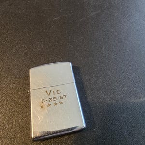 May include: A silver-colored lighter with the name "Vic" and the date "5-29-67" engraved on it, along with four stars. The lighter has a rectangular shape with rounded edges and a hinged lid.