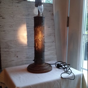 May include: A vintage lamp with a cylindrical base made of cast iron. The base is decorated with a floral design and the letters "F.M.O.". The lamp has a brown cord and a white light bulb.