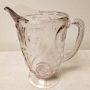 May include: A clear glass pitcher with a decorative pattern and a handle. The pitcher has a wide base and a narrow spout.