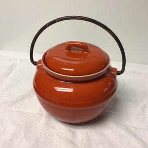 May include: A round, orange-red ceramic cooking pot with a matching lid and a black metal handle. The pot has a white rim and a small handle on the lid. The pot is designed for cooking and serving.