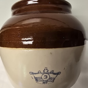 May include: A brown and cream-colored stoneware crock with a dark brown rim. The crock features a blue crown emblem with the number "3" on the front. This vintage piece is likely used for food storage.