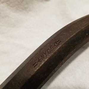 May include: Close-up of a rusty metal bar with the inscription "=NYCS=." The bar is slightly curved and has a hexagonal cross-section.